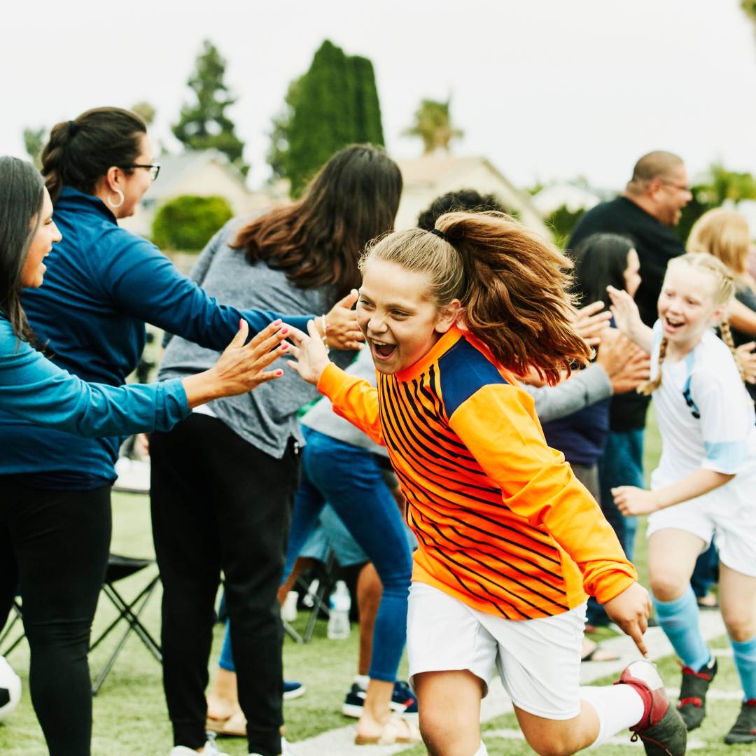 Girls in football kit high-five supporting adults beside a sports field.